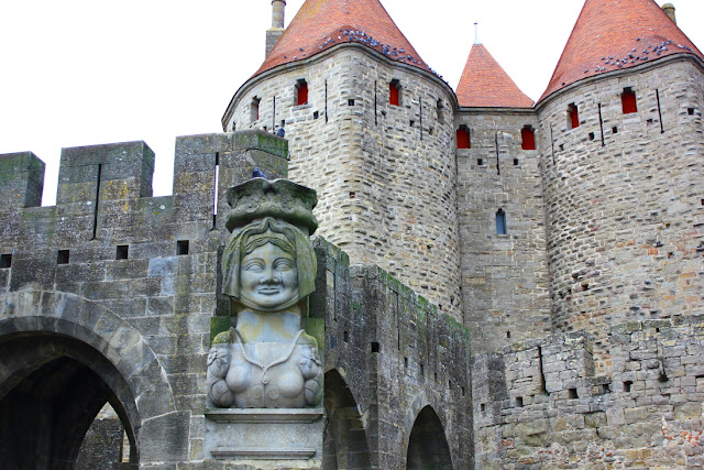 Płaskorzeźba z przedstawienie legendarnej Damy Carsas. W tle widoczny jest fragment średniowiecznej Bramy Narbońskiej z wejściem zamkniętym ostrym łukiem. Ponad krenelażami bramy wnoszą się wieże o dachach krytych czerwoną dachówką.