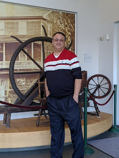 Man wearing blue pants and a blue shirt with red and white stripes. He is standing in front of a museum exhibit that includes a large black and white photo of a house, a spinning wheel, and a larger wheel on a frame