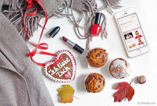 This picture shows a Wiesn themed Flatlay with some dark lipsticks, red nailpolishes, cupcakes and a mobile phone showing our Pinterest board about the perfect Wiesn hairstyles.