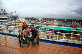 picture of shannon in her manual wheelchair and her daughter on her lap and her husband next to them with a view of the ship and the city skyline in the background