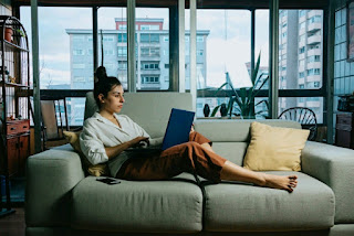 A woman using a laptop while lying on an extended sofa.