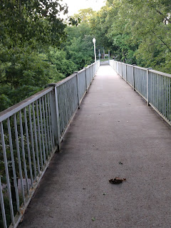 The long concrete Meredith Wilson footbridge with white painted steel railings extends across a creekbed surrounded by trees in Mason City, Iowa