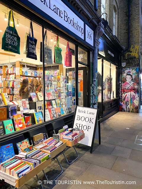 The mural of a woman in colourful clothing on a red brick facade, next to small tables of books which stand in front of an illuminated window of a bookshop decorated with books and cotton tote bags in different colours sporting the name of the shop 'Brick Lane BOOKSHOP' on its front.