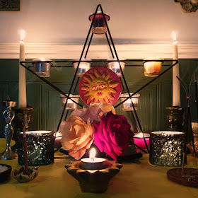 The wall behind is white with white dado rail, then green painted paneling. The altar has two white soy candles in green-glazed chunky candle-sticks. In the middle there is a pentacle candle holder with a purple soy votive candle at the top, a blue soy votive candle at the upper left point, a yellow soy votive candle at the upper right, a green soy votive candle in the bottom left, and a red soy votive in the bottom right, and there five tea-lights in the middle of the pentagram holder at the junctions. A sun plaque in red and yellow is hanging off it. There is a bouquet of mixed roses, peach pink and red, in the middle of the altar. On each side of the roses there is a green candle-jar, mottled with a leaf-shaped tag hanging off it. On the left of the altar there is a Goddess shaped metal incense sconce, on the right there is a pentacle-shaped flat incense burner, three incense sticks are visible. The altar cloth is bright yellow. In the foreground there is a sun-shaped ceramic candle holder with gold leaf and a tea-light. The image has a warm ambiance and was taken in the evening.