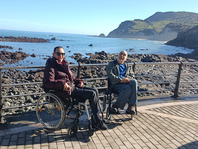 Dos hombres sentados en un paseo, con la costa detrás, un día soleado