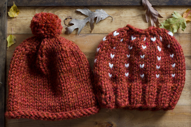 Two rust chunky yarn hats next to each other on a wooden surface. Left hat has a rolled brim and pom pom on top. Right hat has a ribbed brim and cream accent hearts and a hole on top.