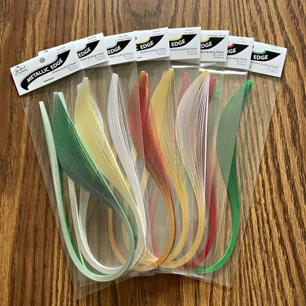 eight packages of metallic edge quilling strips in a variety of holiday colors displayed on table.