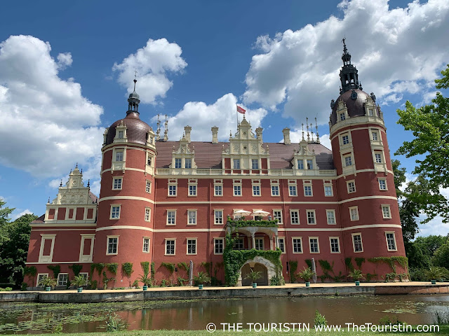 Water lilies on a lake in front of a red-ish coloured renaissance castle with an overgrown balcony under a blue sky