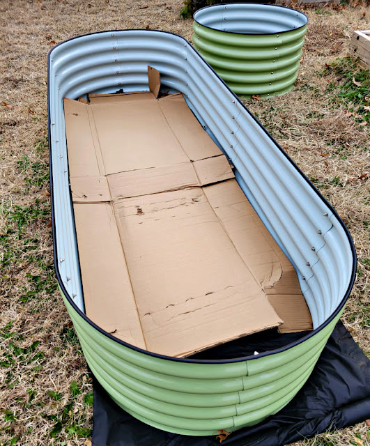 Finished, empty metal raised garden beds, one oval and one round, with cardboard lining the bottom.