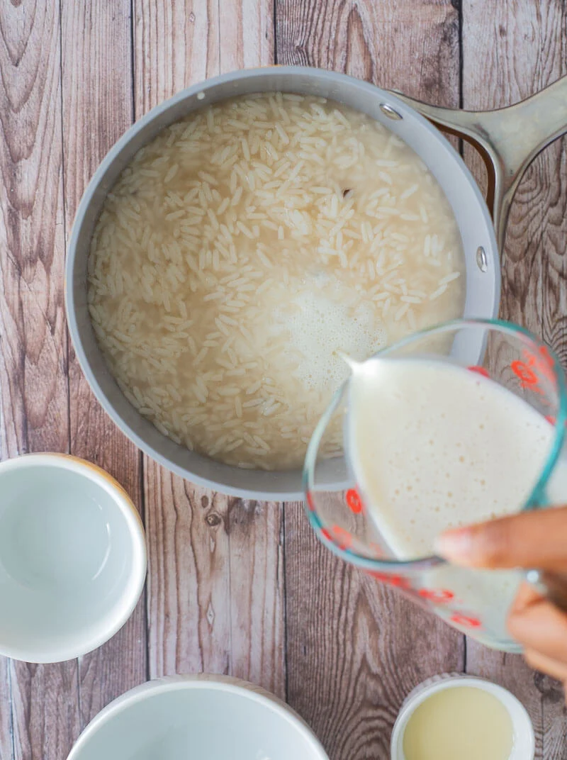 A measuring cup of milk being poured into the boiled rice with water and spices to make the porridge creamy.