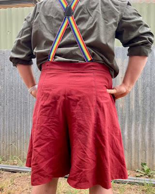 The back of a pair of red culottes, worn by a white person. They are attached to rainbow suspenders. End ID.