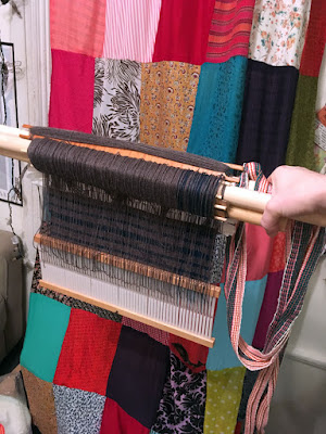 A white hand holding a pair of dowels with a frankly alarming amount of yarn wrapped around them and hanging down from them, suspending a rigid heddle and showing a horizontally striped grey and teal piece of cloth behind the threads, against a bright patchwork curtain.