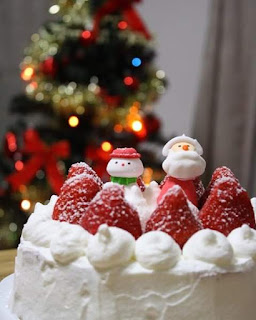 Foto de um bolo branco. Merengues decoram a borda. Ao centro e cercados por morangos açucarados, dois enfeites lado a lado, um papai noel e um boneco de neve. Ao fundo, em desfoque, uma árvore de Natal com luzes coloridas, laços vermelhos grandes e outros enfeites.  Desejamos um Natal iluminado para todos!