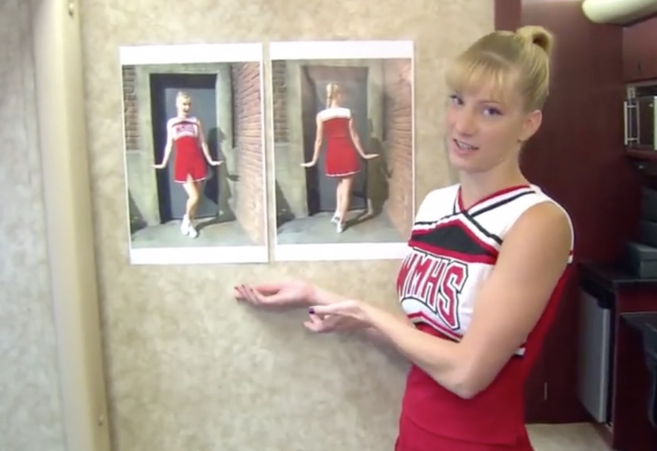 Heather Morris as Brittany, a young woman in red and white high-school cheerleader uniform with blond hair pulled up into ponytail gesturing at photos of herself in that same outfit on a wall