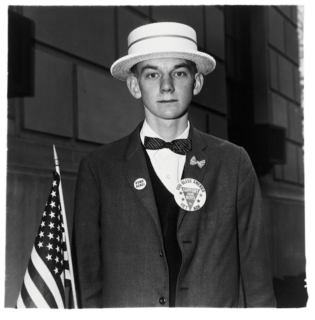 Foto em preto e branco, de um menino sério, usando terno com alguns broches, segurando uma bandeira dos Estados Unidos, e usando um chapéu feito de canudos