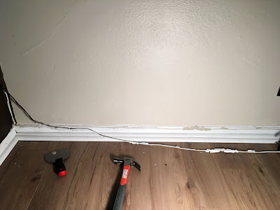 A bad indoors-at-night photo of a wire with large flakes of paint stuck to it, hanging where it was pulled from just above the baseboard against a cream wall, with a hammer and a small metal joint knife on the floor.