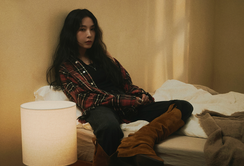 A promotional shot of BoA for her single “Emptiness”. Featuring her sat on a bed, wearing a red check flannel shirt, jeans and brown suede boots.