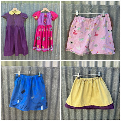 A photo collage of four different photos. Top left shows two dresses side by side. The left is dark purple with a yellow peter pan collar, puffed short sleeves, and an empire waistband. It buttons up the front with three large, bronze-look, heart-shaped buttons. The dress on the right has a bodice of purple quilting cotton with small circles in darker purple. The sleeves and skirt are made of bright pink fabric with equally spaced, flower-like mandala patterns. The dress has an elasticated peasant blouse neckline, and the short sleeves are puffed. There is a ruffle around the hem made of the purple fabric, and a ruffle made of the pink fabric placed vertically down the front of the bodice.   The bottom left photo shows a child's gathered skirt made of shiny blue material. There are small lengths of ribbon, in several different shades of blue, stitched down randomly over the fabric.  The bottom right photo shows a pair of child's shorts made of light pink fabric covered in cute animal motifs. They have brown stripes down the side.