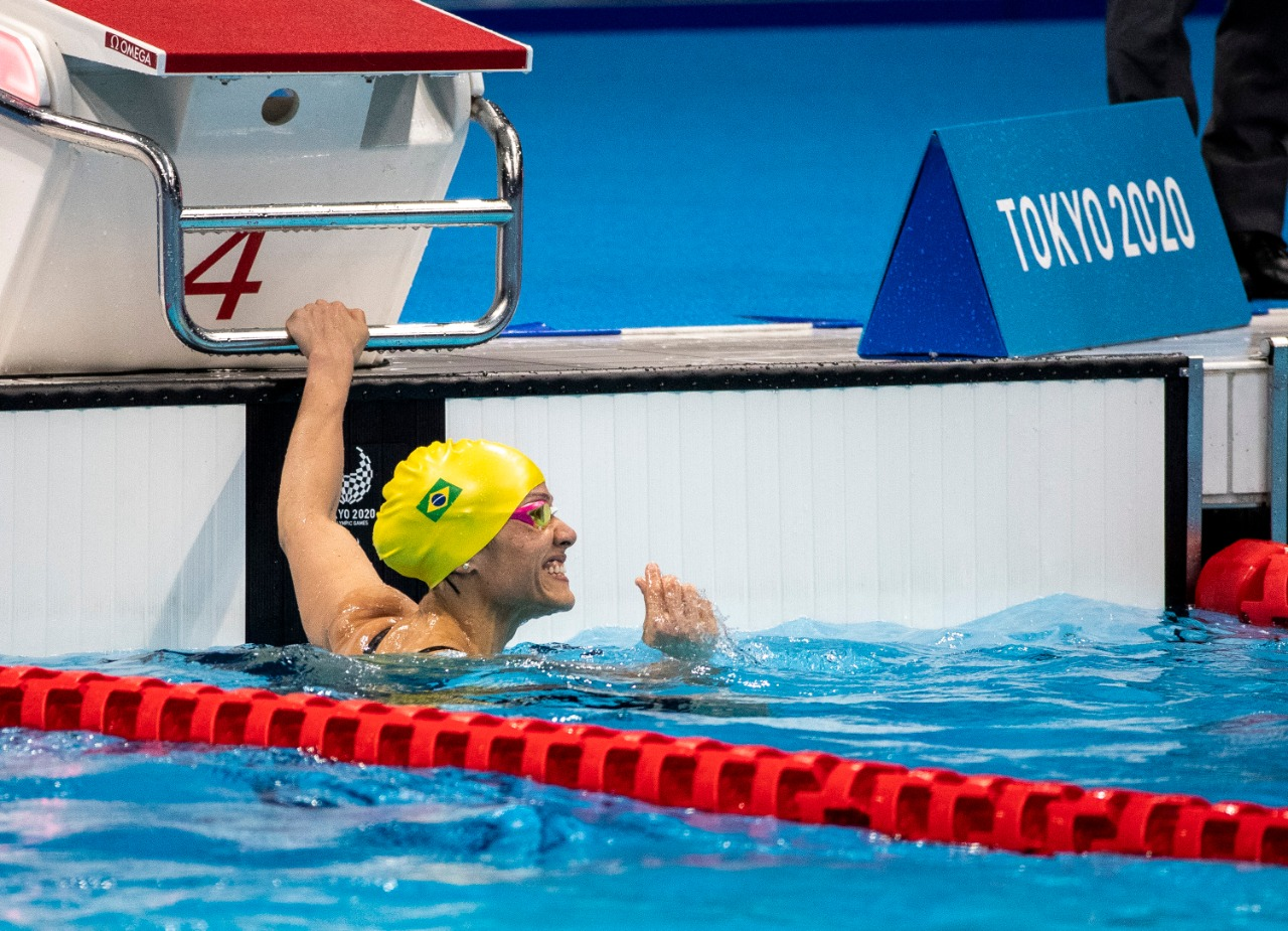 De touca amarela e óculos rosa, Carol Santiago sorri segurando na borda da piscina