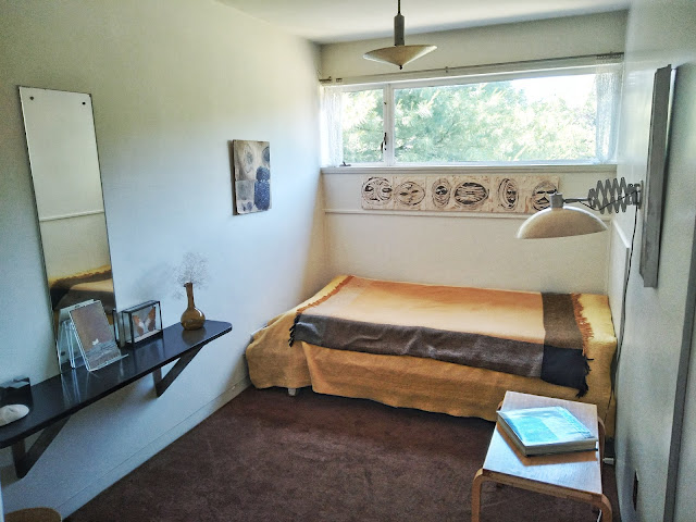 A wide angle view of a bedroom, with a thin mirror and shelf holding knickknacks to the left, a daybed with a yellow cover from center to left. Abstract art hangs between the daybed and a wall length window.