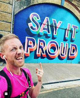 House of Happiness event manager, Neil, is depicted in front of a brightly coloured and lettered outdoor sign which reads 'Say It Proud'