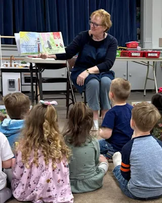 woman with short, blonde hair holds open book while showing collage art illustrations to schoolchildren seated on floor in classroom