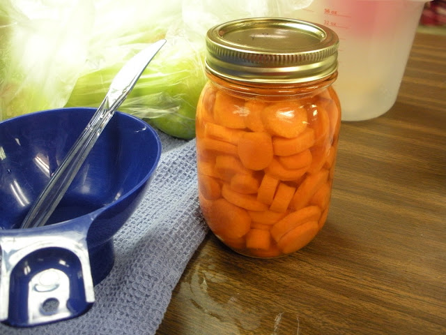 A pint jar of pressure-canned carrots with canning equipment including a blue plastic funnel.