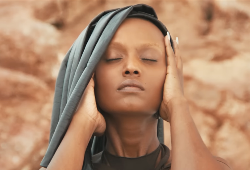 A shot of Kelela in her music video for “Washed Away”, shot in close up, about to remove her hat / headdress which mimics the hair style she had during the release of ‘Take Me Apart’..