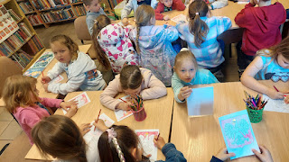 sala biblioteczna. Grupa przedszkolaków oraz dwie panie przedszkolanki siedzą przy bezowych drewnianych stolikach. dzieci kolorują kolorowanki przedstawiające herb Polski. W tle widać regały z książkami.