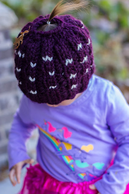 Toddler wearing a lavender top with a fuchsia tutu is looking down so that the tiny straight up pony tail of fine hair is visible poking through a purposely made hole on top of a knitted dark purple beanie with cream accents. Dried oak leaf stuck on hat.