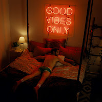 man lying on a bed with a neon sign above saying “Good vibes only”