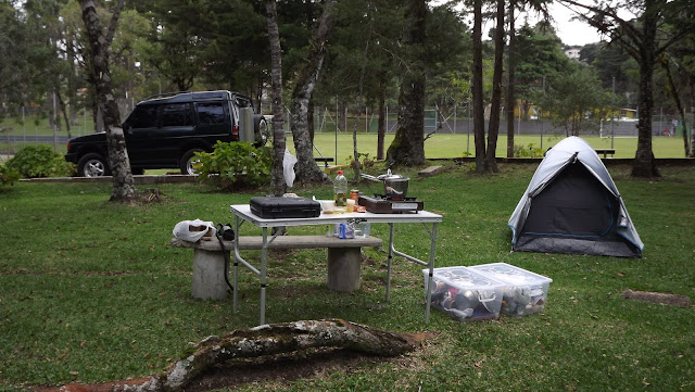 Nossa área de camping, com a nossa Malandra (Discovery 1 300tdi de 1996 verde) ao fundo, nossa barraca (Quechua Arpenaz 2 Fresh and Black) sobre a grama ao lado de uma mesa dobrável com coisas de camping.