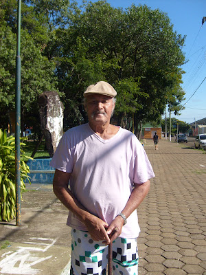 Um homem enquadrado dos joelhos até acima da cabeça, usando boina, camiseta, bermuda e segurando uma bengala de madeira.