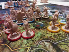 A group of beige figurines and one pale blue-grey figurine, all resembling the stereotypical image of Vikings, with red, yellow, or dark brown bases to differentiate player factions, in the central space of the game board. The board represents an island divided into various provinces, painted to look like generic Nordic terrain. A square token can be seen in each province with one of three symbols: a hand holding lightning bolts, an axe head, and a stereotypical Viking helmet complete with anachronistic horns. The tile in the center province contains all three of these symbols.