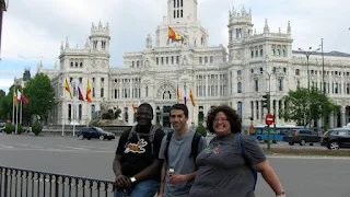 Three people standing in front of a fence that separates the sidewalk form a busy multi-lane street. A large white building is in the background. The building is decorated in a gothic style and looks almost like a castle with many Spanish flags hanging from it.