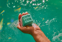 A wide angled phot of a square cube bath bomb filled with grains of epsom salt with lush CBD engraved into the top dissolving and letting out blue colours into a tub of water on a bright background