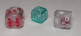 On the left is a large, hollow, clear plastic d6 with red pips, inside of which are two tiny red d6s with white pips. Beside that is a clear green plastic d6 with white pips, slightly larger than a normal d6, that is also hollow, and inside is a tiny white d6. On the right is a clear hollow d10 with white numbers, which has a small red d10 inside of it.