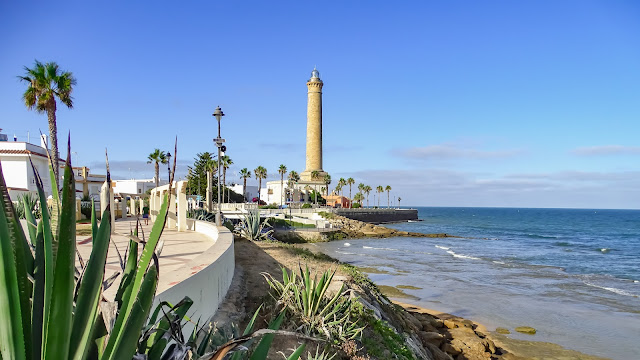 Paseo marítimo y pequeña playa rocosa con una gran chimenea industrial al fondo.