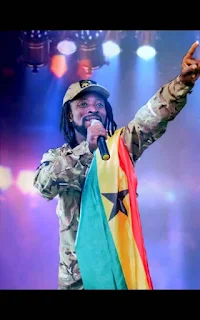 Bongofari in a military attire with a Ghana flag hanging on his left shoulder a mic on his right sing on stage.