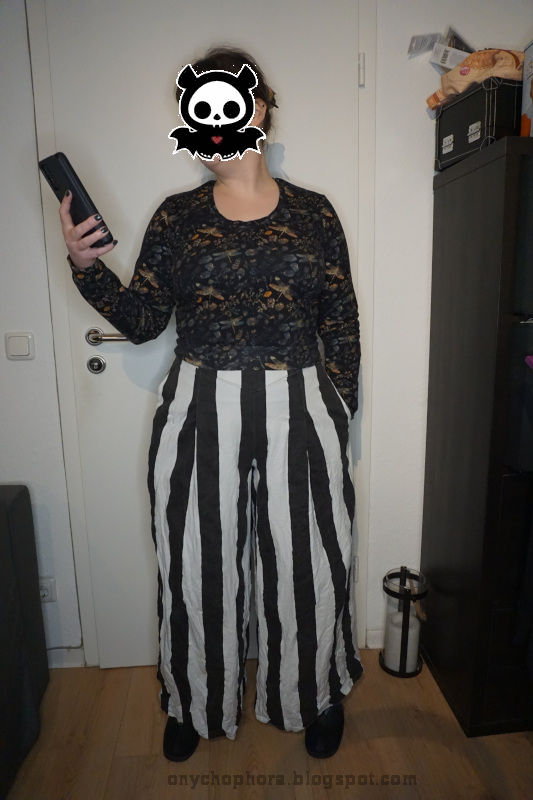 Frontalansicht von einer stehenden Frau. Sie trätgt eine schwarz-weiß längsgestreifte, weite Hose und einen dunklen Pullover mit Libellen- und Blumenmuster. Der Pullover ist nicht in den Bund gesteckt, sodass man die Länge sieht.