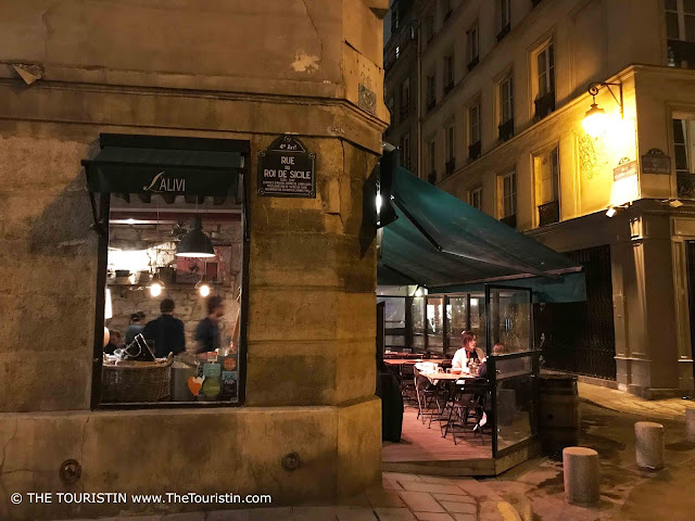 View through a window into a restaurant in Marais with a blue street sign and a green awning on its facade.