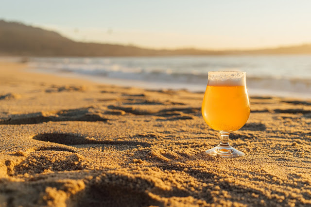 Un bicchiere di birra bionda sulla destra dell'immagine lasciato su una spiaggia di sabbia dorata al tramonto e circondato da impronte
