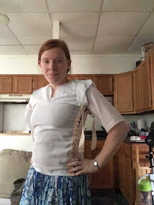 A young, redheaded white womanstanding diagonally to the camera with her left hand on her hip, with wooden kitchen cabinets behind her, wearing silver bracelets on either wrist and an extremely snug palest aqua bodice, with a short neck slit and laced shut through tapes at the front and back sides with pale blue ribbon. A small cp sleeve of excess fabric has formed on top of her shoulder, and a close-fitted half sleeve ends at her crooked elbow. The too-long ends of the lacing tapes dangle from just under her armpit.