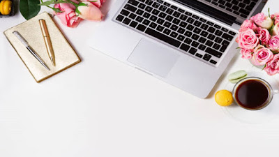 Two Gold and silver pens on a white notepad, red rose flowers, computer and a glass of beverage on top of a white work table.