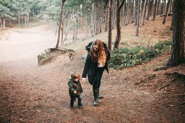 image shows a mother and toddler son walking through the woods in autumn. They are dressed in warm clothes.