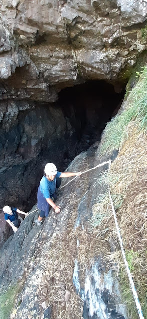 Photograph from above of two women in helmets climbing down a steep rocky slope into a cave