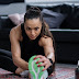 Woman in black tank top sitting on a floor doing arms and legs stretching at home