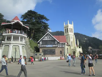 Visitors walking around The Ridge in Shimla, with the historic Christ Church, Shimla State Library, and a scenic hill backdrop under a bright blue sky.