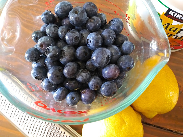 a cup full of fresh blueberries next to two fresh lemons and a bottle of pure maple syrup