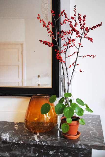 Op een marmeren mantel staat een okergeel glazen vaasje, een doorzichtige vaas met takken en een klein Pilea plantje.